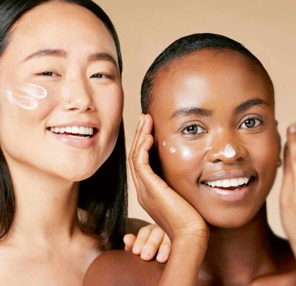 Zwei glückliche Frauen haben auf Nase und Wangen Hautpflegecreme vom Online-Shop für kosmetische Pflegeprodukte aufgetragen. Sie haben eine reine und jung wirkende Gesichtshaut. Kosmetikerin, ihre Hand zu sehen, flüstert einen der Frauen etwas ins Ohr.