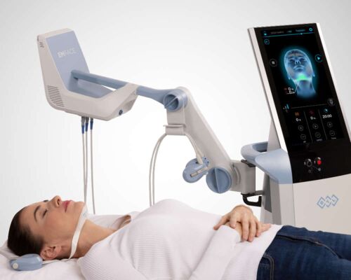 Kundin liegt entspannt auf einer Liege während einer Behandlung des Doppelkinns mit EMFACE® Submentum. Gerät mit der Technologie zur Kinnverschönerung durch Fettreduktion und Hautstraffung steht daneben. Am Hals der Frau ist ein Applikator angebracht.