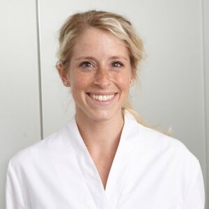 Eva Klütsch ist in einen weißen Arbeitskittel gekleidet. Ihre blonden Haare sind zu einem Zopf gebunden. Sie lächelt und ihre Augen wirken glücklich. Frau Klütsch ist eine staatlich anerkannte Podologin und Spezialistin für schöne und gesunde Füße.