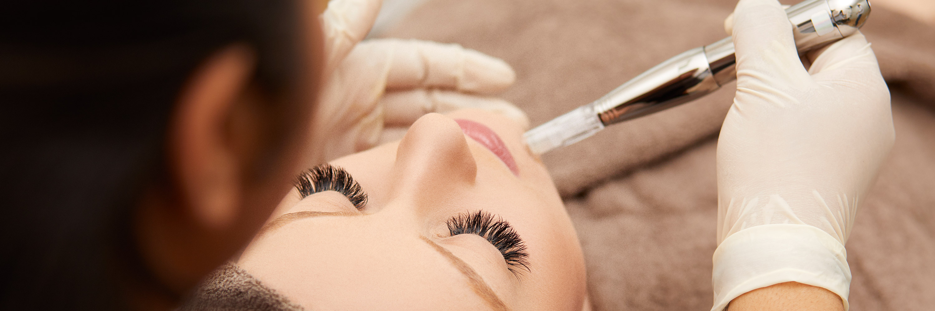 Während einer medizinischen Microneedling-Behandlung liegt eine Patientin mit geschlossenen Augen in Handtücher gehüllt auf einer Behandlungsliege. Eine Kosmetikerin fügt mit dem Dermapen kleine Stiche der Gesichtshaut der Kundin zu.