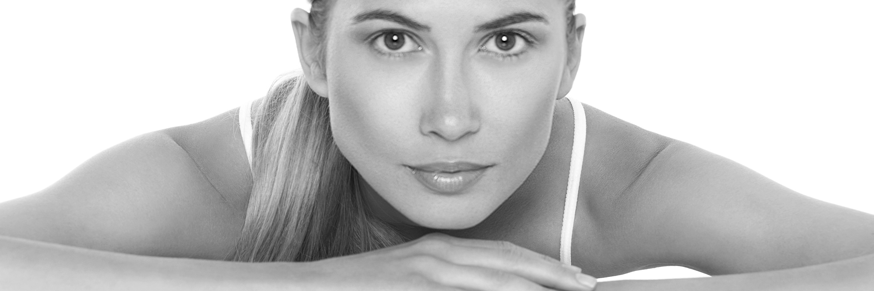 Aus einer s/w Fotografie blicken leuchtende Augen einer jungen Frau auf den Betrachter. Ihr Gesicht enthält nach einer Hautbehandlung mit Venus Viva keine Falten oder Narben und wirkt makellos. Sie hat ihre Arme angewinkelt auf einen Tisch gelegt.