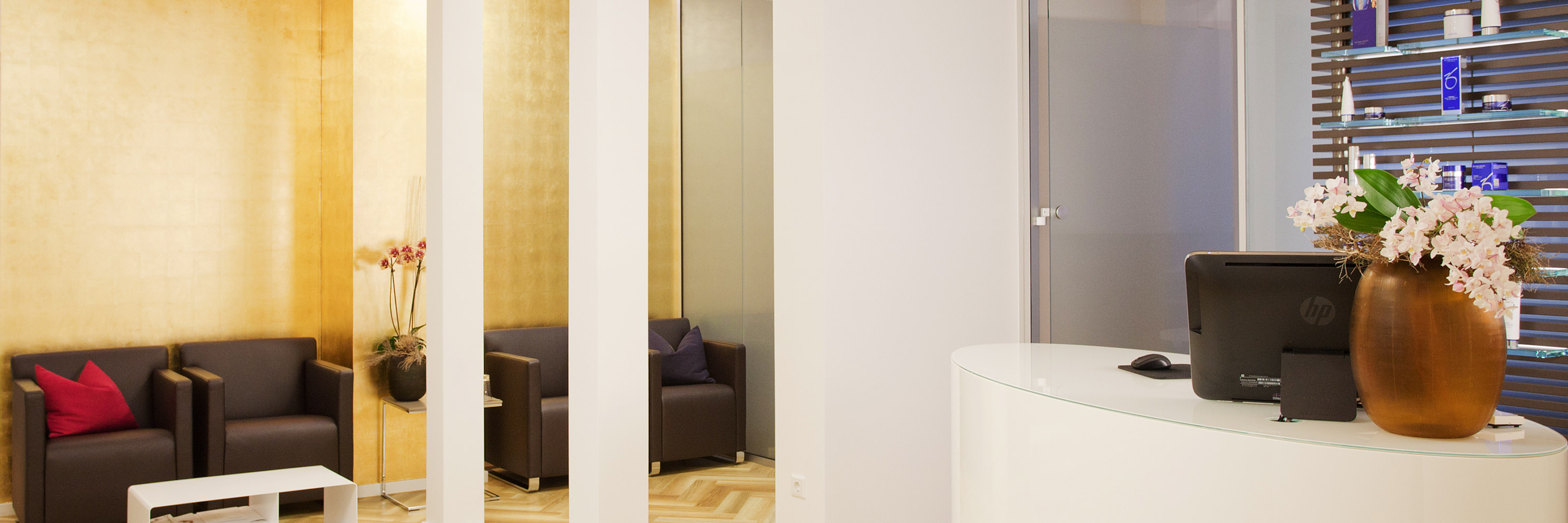 Empfangsraum des Instituts für Hautpflege und Hautbehandlungen in Frankfurt am Main. Im Vordergrund eine Empfangstheke mit frischen Blumen und Monitor. Im Hintergrund dunkle weiche Sessel. Die Wand hinter den Sesseln ist in Gold.